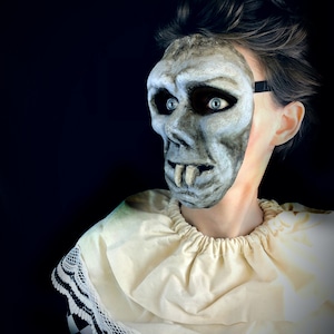 May include: A person wearing a white and black checkered costume with a white lace collar and a creepy, gray, realistic-looking mask with a wide, toothy grin.