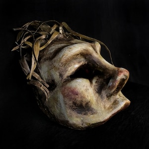 May include: A papier-mâché mask with a distressed, textured finish. The mask is painted in shades of brown, beige, and red. It has a crown of woven straw or twigs.
