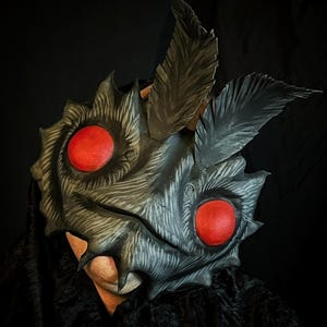 Könnte beinhalten: Eine von einer Motte inspirierte Maske mit einer dunkelgrauen, strukturierten Oberfläche und feinen, geätzten Linien, die Fell imitieren. Die Maske hat zwei markante, kreisförmige, leuchtend rote Augen und zwei große, spitze, federartige Antennen.