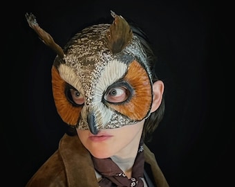 Puede incluir: Una máscara de búho detallada con colores marrón, naranja y blanco. La máscara presenta ojos grandes y expresivos y estructuras puntiagudas en forma de orejas. La persona lleva una chaqueta de ante marrón y un pañuelo marrón.