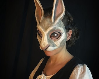 Puede incluir: Una máscara de conejo gris y blanco con largas orejas, pintada con sutiles sombras. La máscara se lleva sobre la cara, con los ojos del usuario visibles. La persona lleva un chaleco negro sobre una blusa blanca.