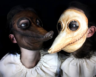 May include: Two people wearing plague doctor masks. One mask is brown with a stitched beak, the other is off-white with a long beak. Both masks have large eye openings and are set against a black background.