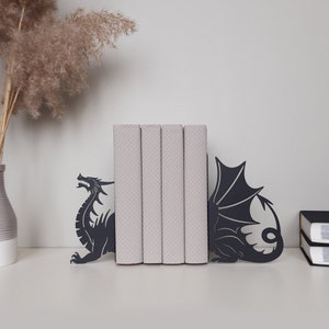 K&ouml;nnte beinhalten: Schwarze Metall-Drachen-Buchst&uuml;tzen, die einen Stapel B&uuml;cher mit gemusterten Einb&auml;nden halten. Die Buchst&uuml;tzen sind in Form von Drachen mit Fl&uuml;geln und Schw&auml;nzen gestaltet.