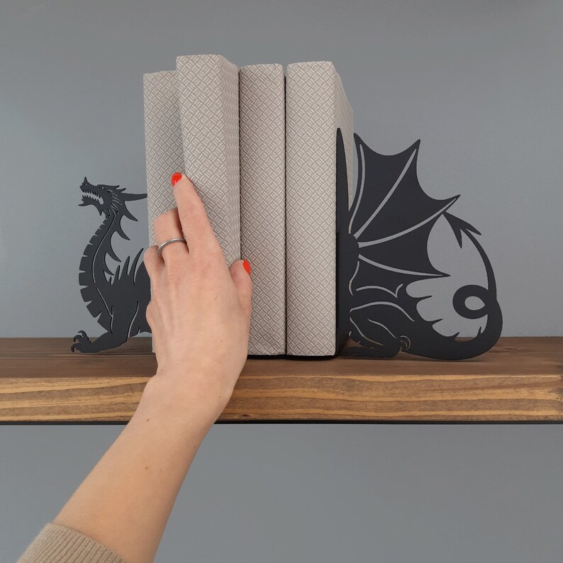 K&ouml;nnte beinhalten: Schwarze Metall-Drachen-Buchst&uuml;tzen, die drei B&uuml;cher mit gemustertem Einband halten. Die Buchst&uuml;tzen stehen auf einem Holzregal.