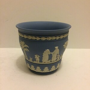 May include: A small, blue ceramic planter with a white design of people and palm trees. The planter has a scalloped rim and a white base.