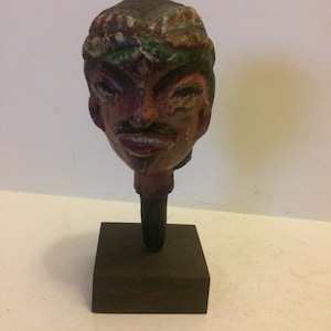 May include: A hand-painted wooden puppet head with a colorful turban, a red face, and a mischievous expression. The head is mounted on a wooden base.