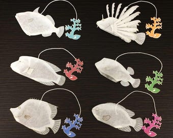 May include: A collection of six white fish-shaped tea bags with colorful coral accents. Each tea bag has a white string attached. The coral accents are in shades of blue, orange, red, green, and pink. The background is a dark, textured surface.