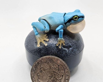 Könnte beinhalten: Eine kleine blaue Froschfigur mit grünen Augen und einem weißen Mund, die auf einem runden schwarzen und grauen Sockel sitzt. Die Figur ist etwa so groß wie ein Silberdollar.