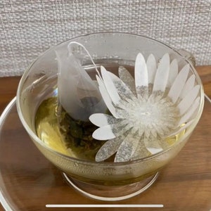 May include: A clear glass teacup and saucer with a daisy design, filled with green tea and a tea bag. The cup sits on a wooden surface, with a neutral-colored background.
