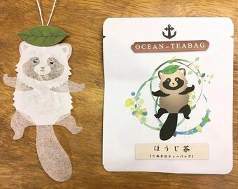 May include: A white tea bag with a cartoon raccoon design. The tea bag is shaped like a raccoon with a leaf on its head. The packaging is white with blue and green watercolor designs and the text "Ocean-Teabag" and "ほうじ茶 [ほうじ茶ティーバッグ]" in Japanese.