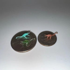 May include: Three miniature plastic dinosaur toys in teal, green, and peach colors. The dinosaurs are placed on top of two coins. The dinosaurs are small, with the largest being about 1 inch in length.