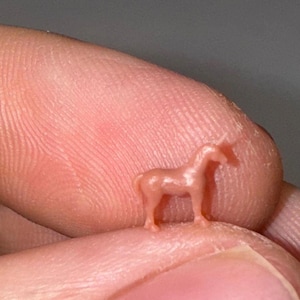 May include: A tiny, light pink unicorn figurine. The miniature horse-like creature is standing upright, with a slender body and a pointed horn. The figurine is held between two fingers.