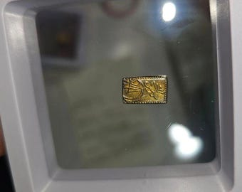 May include: A small, gold-colored, rectangular metal object with intricate designs. The object is framed in a clear plastic case.