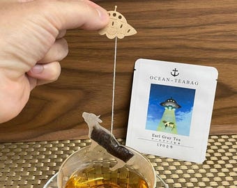 May include: A tea bag with a brown UFO design hanging from a string. The tea bag is in a glass cup with tea. The tea bag packaging is white with blue text that reads "OCEAN-TEABAG" and "Earl Gray Tea UFO24".