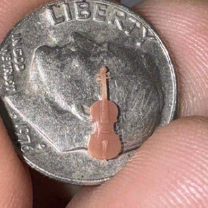 May include: A tiny, light brown violin figurine rests on a silver dime. The miniature instrument is detailed, with visible strings and a bow. The dime's surface shows the word "LIBERTY" and the year 1976.