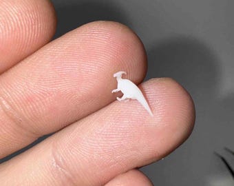 May include: A tiny, white Parasaurolophus dinosaur figurine. The miniature sculpture is held between fingers, showcasing its detailed form, including the distinctive crest and tail. The figurine is a pale, solid color, and appears to be made of plastic or resin.