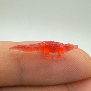 May include: A small, translucent red alligator figurine. The miniature reptile is detailed with textured skin and tiny legs, appearing to be made of a clear, reddish material. The figurine is resting on a fingertip.