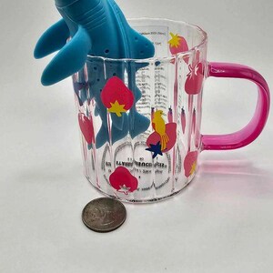May include: A clear glass mug with a pink handle and a strawberry and bird design. A blue silicone shark tea infuser is in the mug.