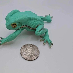 May include: A green frog figurine with a gold eye, sitting on a white surface next to a US quarter.