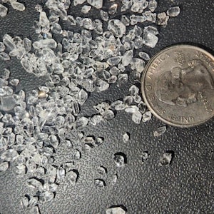 May include: A close-up image of a pile of small, clear quartz crystals on a black surface. A US quarter is included for scale.
