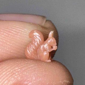 May include: A miniature, light pink squirrel figurine, posed in a seated position while holding a small object. The squirrel has a fluffy tail and is detailed with textured fur. The figurine is small, likely under 1 inch in height.
