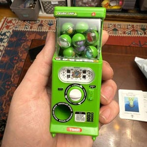 May include: A green toy vending machine with the text "COSMIC CODE" and "TWIZZLER SPACE". The machine is filled with green and clear plastic balls. The machine is held in a hand.