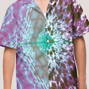 May include: Short-sleeved button-down shirt with a vibrant, abstract geometric design in shades of purple, teal, and brown. The shirt features a collared neckline and a split design, creating a unique visual effect. The shirt is paired with light blue shorts.