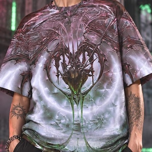 May include: A short-sleeve t-shirt featuring an intricate, symmetrical graphic print in shades of purple, green, and white. The design displays abstract, organic, and sharp-edged patterns across the front. The shirt has a crew neckline and a loose fit.