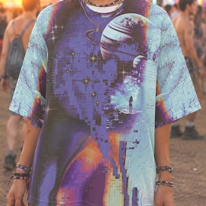 Könnte beinhalten: Ein kurzärmeliges T-Shirt mit einem lebendigen kosmischen Grafikdruck, der einen großen Planeten, Sterne und die Silhouette einer Figur auf einem Felsvorsprung zeigt. Das Design kombiniert tiefe Violett-, Blau- und Orangetöne mit einem Rastermuster.