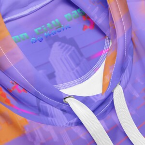 May include: A close-up of a purple hoodie with a cityscape design. The hoodie features a drawstring and a white interior. The design includes the text "Neon City Daze by Kadix" in a pixelated font, with orange and pink accents.
