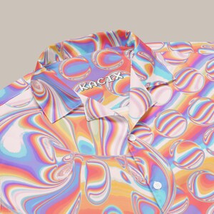 May include: A short-sleeved shirt with a vibrant, psychedelic pattern in shades of orange, pink, purple, blue, and yellow. The shirt features a camp collar and a button-down front. The brand name "KACIX" is visible on the collar.
