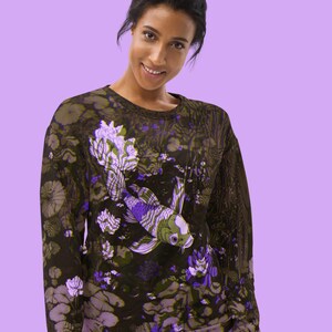 Puede incluir: Una sudadera negra con un estampado floral morado y verde que presenta un pez koi. El pez nada en un estanque con nen&uacute;fares y otras plantas acu&aacute;ticas.