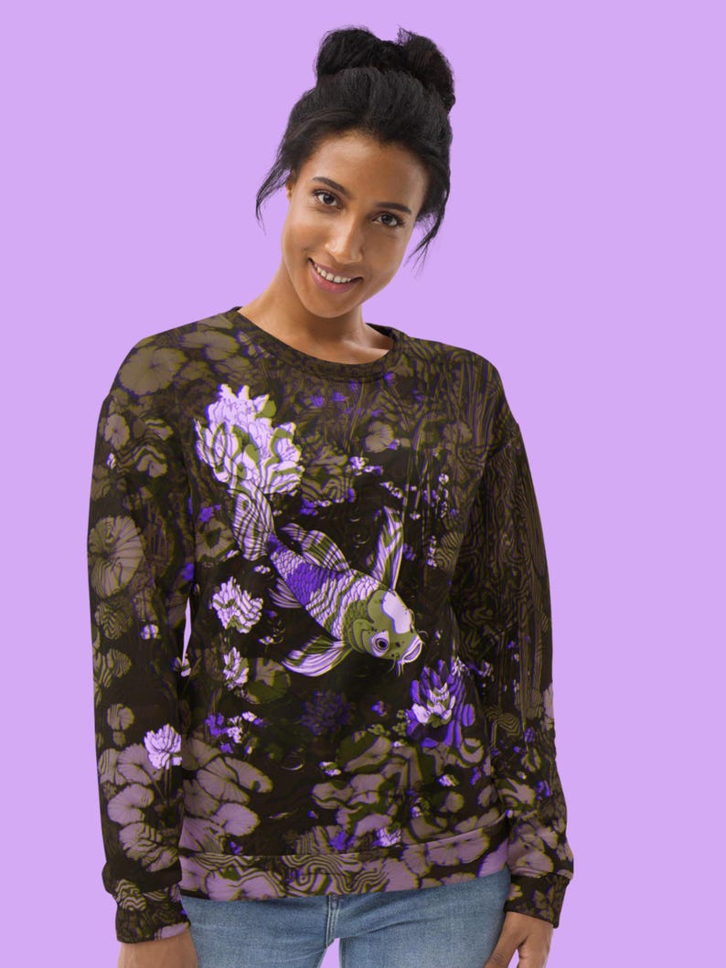 Puede incluir: Una sudadera negra con un estampado floral morado y verde que presenta un pez koi. El pez nada en un estanque con nen&uacute;fares y otras plantas acu&aacute;ticas.