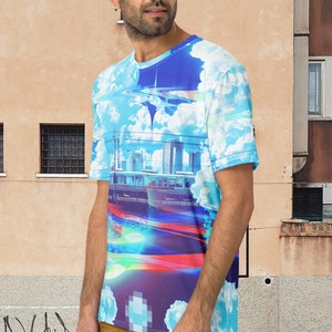 Op de afbeelding: Een blauw, wit en paars T-shirt met een grafische print van een stadsbeeld en wolken. Het shirt heeft een ronde hals en korte mouwen.
