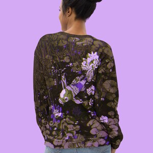 Puede incluir: Una sudadera negra con un estampado floral morado y verde con un pez koi blanco y morado. La sudadera tiene mangas largas y cuello redondo.