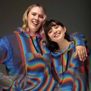 May include: Two people wearing colourful, abstract print hoodies. The hoodies have a black drawstring and a kangaroo pocket. The people are smiling and looking at the camera.