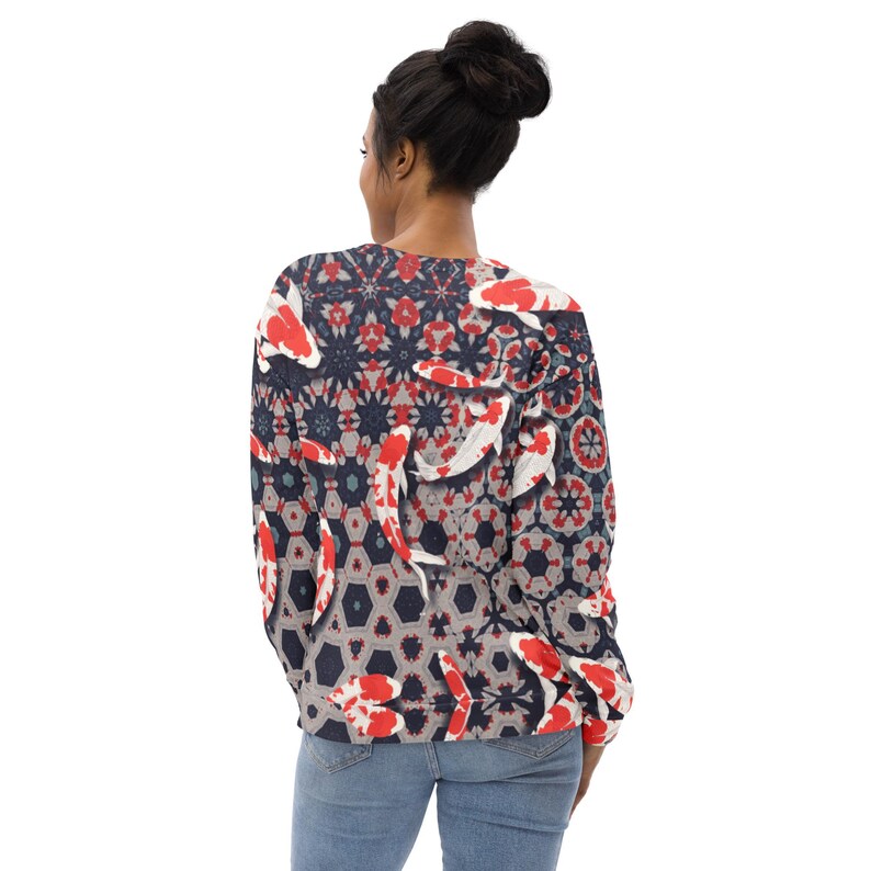 May include: A long-sleeved sweatshirt featuring a koi fish pattern. The design incorporates red, white, and black koi fish against a geometric background of gray, navy, and red shapes. The sweatshirt is worn by a person wearing blue jeans.