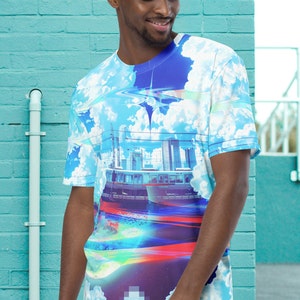 Op de afbeelding: Een blauw, wit en rood t-shirt met een stadsbeeld en wolken erop gedrukt. Het shirt heeft een ronde hals en korte mouwen.
