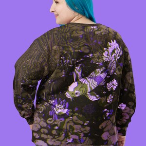 Puede incluir: Una sudadera negra con un estampado floral morado y verde que presenta un gran pez koi. El pez nada en un estanque con nen&uacute;fares y otras plantas acu&aacute;ticas.