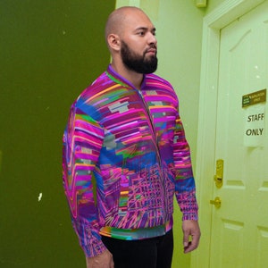 Puede incluir: Un hombre lleva una chaqueta bomber colorida con un patr&oacute;n geom&eacute;trico. La chaqueta tiene una cremallera y mangas largas. El hombre est&aacute; de pie frente a una puerta con un cartel de "Solo personal".