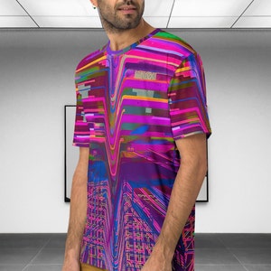 May include: A man wearing a short-sleeved t-shirt with a colorful abstract design. The design features a mix of pink, purple, blue, and yellow lines and shapes.