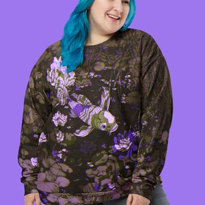 Puede incluir: Una sudadera negra con un estampado floral morado y verde que presenta un pez koi. El pez nada en un estanque con nen&uacute;fares y flores.