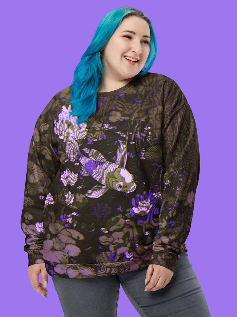 Puede incluir: Una sudadera negra con un estampado floral morado y verde que presenta un pez koi. El pez nada en un estanque con nen&uacute;fares y flores.