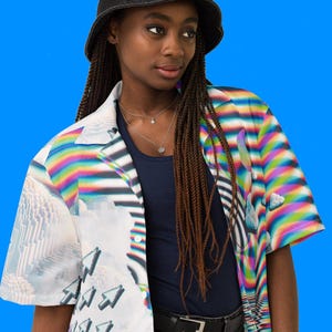 Puede incluir: Una persona lleva una camisa blanca abotonada con un dise&ntilde;o colorido, pixelado y abstracto. La camisa tiene mangas cortas y cuello. La persona tambi&eacute;n lleva un sombrero de pescador negro y pantalones vaqueros negros.