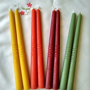 Op de afbeelding: Acht tapende kaarsen in geel, rood en groen met een decoratief patroon. De kaarsen zijn in paren gerangschikt op een wit oppervlak.
