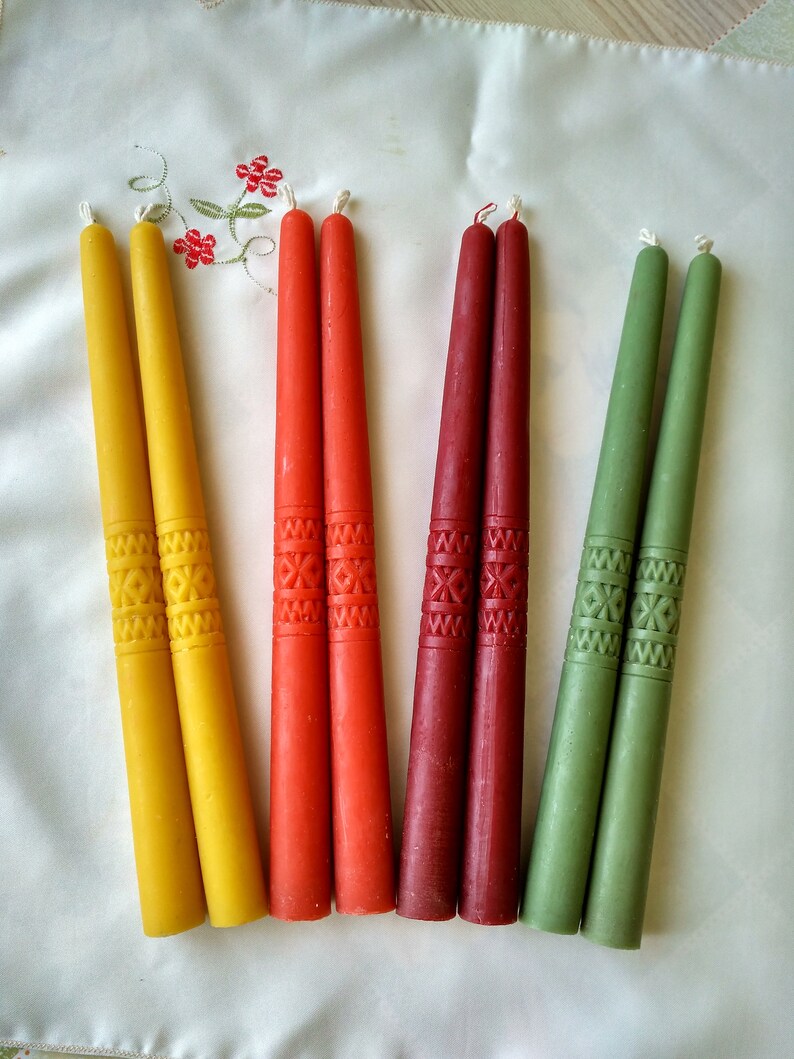 Op de afbeelding: Acht tapende kaarsen in geel, rood en groen met een decoratief patroon. De kaarsen zijn in paren gerangschikt op een wit oppervlak.