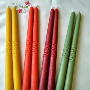 Op de afbeelding: Zes taps-kaarsen in geel, rood en groen. De kaarsen zijn versierd met een herhalend geometrisch patroon.