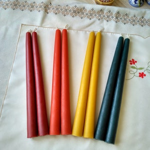 May include: Six taper candles in red, orange, yellow, and dark blue. The candles are arranged in pairs on a white surface.