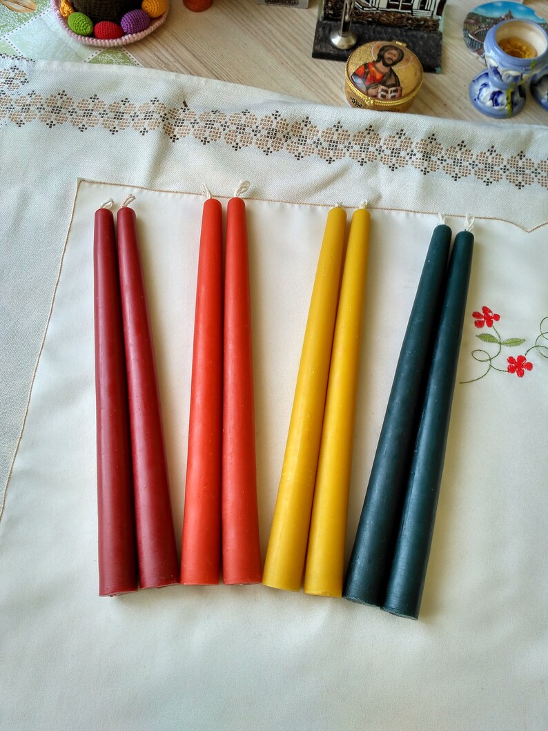 May include: Six taper candles in red, orange, yellow, and dark blue. The candles are arranged in pairs on a white surface.