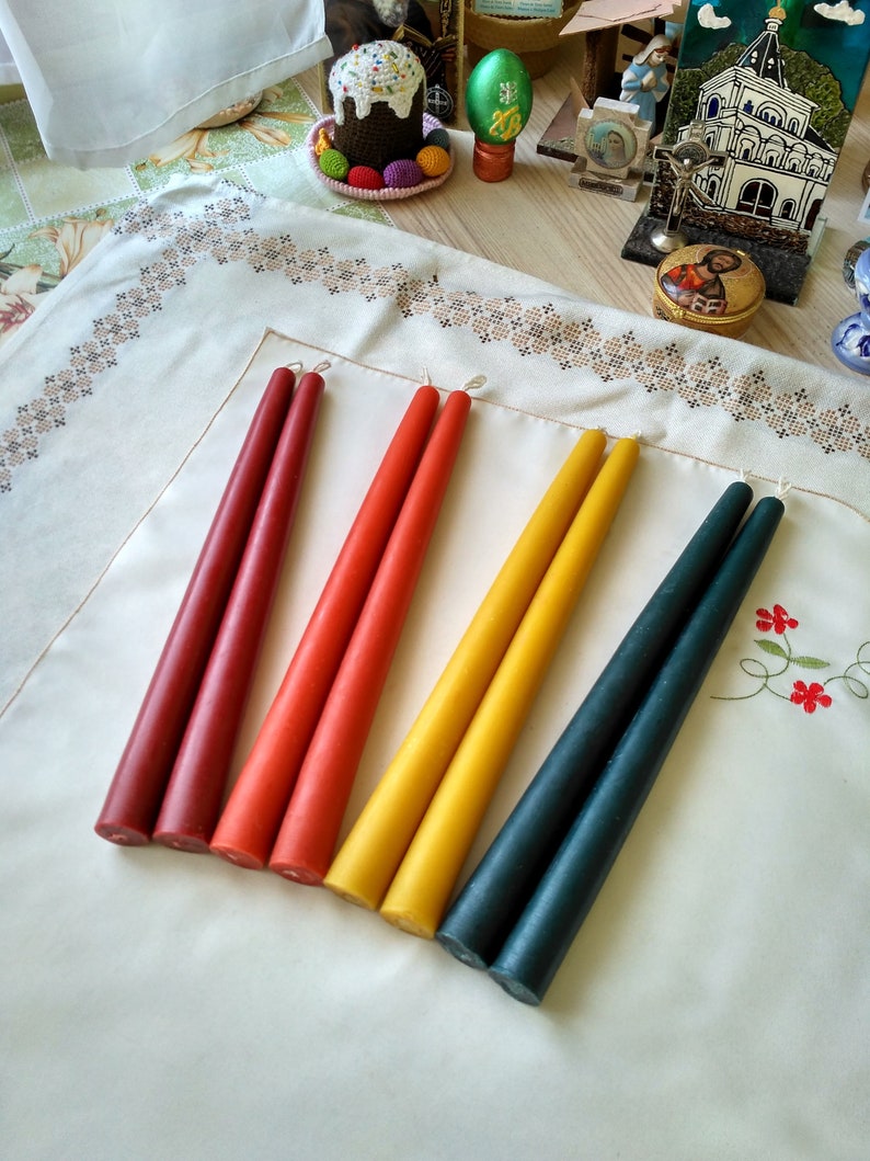 May include: Five tapered candles in various colors, including red, orange, yellow, and green, arranged in a row on a white tablecloth.